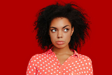 Serious African American woman with natural curly hair, wearing a coral pink polka dot shirt, looking sideways with a skeptical expression against a bold red background..