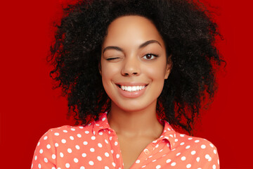 Playful African American woman with natural curly hair, winking and smiling, wearing a coral pink polka dot shirt against a bold red background.