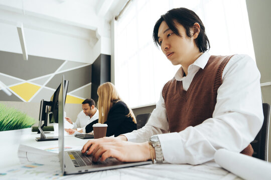 Asian architect working on laptop in modern office with colleagues analyzing blueprints