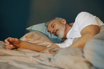 Elderly man lying on his side on comfortable bed, resting his head on pillows and sleeping peacefully with relaxed expression