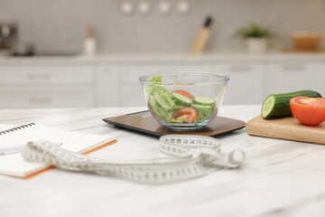 Salad on scales, notebook and measuring tape in kitchen, closeup