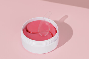 White container jar with pink hydrogel eye patches and a spatula, set against a pink isolated backdrop. Targets moisture retention, fine lines, under-eye puffiness, and elasticity of the skin.