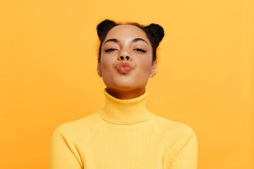 Playful African American woman with natural curly hair styled in two buns, wearing a yellow turtleneck sweater, sending a kiss and smiling with puckered lips against a bright yellow background.