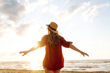 Back view of a young woman with curly hair feels free on the seashore. Young woman walks on the beach at sunset. Concept of relaxation, enjoyment, freedom.