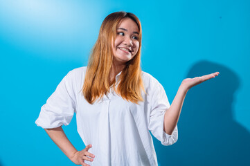 Fototapeta premium Asian woman with long straight hair in white shirt smiling while holding one hand open to the side as if presenting or showing something, standing confidently against a solid blue studio background
