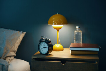 Cozy bedroom nightstand setup showcasing a yellow lamp, an alarm clock, books, and water bottle,...