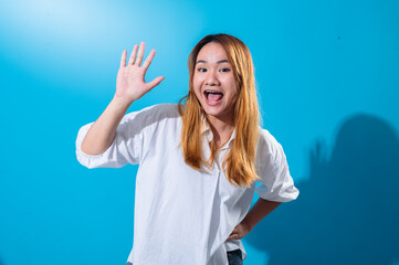Fototapeta premium Asian woman with long straight hair in white shirt smiles cheerfully while waving her hand in a friendly greeting pose, standing against solid bright blue background in a well-lit studio setting