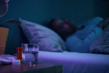 Person lying in bed under soft, blue-hued lighting with a glass of water and pill bottles on a bedside table. Relaxing evening scene with focus on health and rest