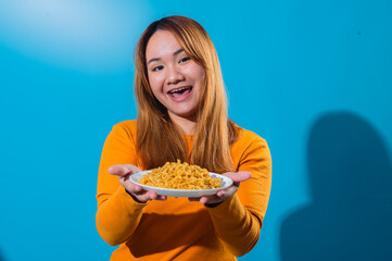 Happy Asian woman in orange shirt smiling and winking while holding a plate of instant noodles with...