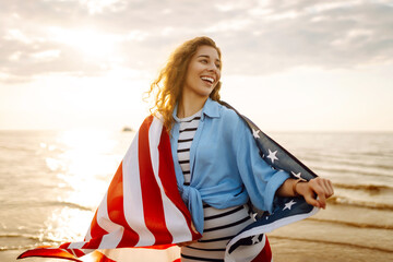 Fototapeta premium Portrait of happy woman with American flag humming on sandy shore at sunset. Young woman feeling freedom enjoying seascape. Independence concept.