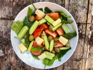A vibrant and healthy vegetable salad served in a white bowl on a rustic wooden surface. The salad includes fresh spinach leaves, cucumber sticks, tomato wedges, green olives, and cubes of hard cheese