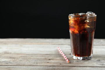 Refreshing cola with ice cubes in glass and straw on wooden table against black background, closeup. Space for text