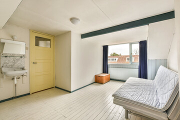 A minimalist interior with light wood flooring, a simple mattress, and a small window providing natural light. The room's design emphasizes simplicity and functionality.
