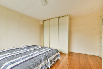 A serene minimalist bedroom featuring a bed with a striped blanket and a large wardrobe. The natural light enhances the warm tones of the wooden floor.