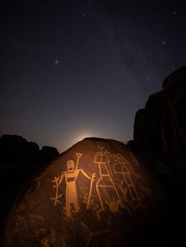 a ancient petroglyphs on a stone wall under a starlit night sky. The illuminated carvings depict human figures and symbols, blending archaeological significance with celestial wonder