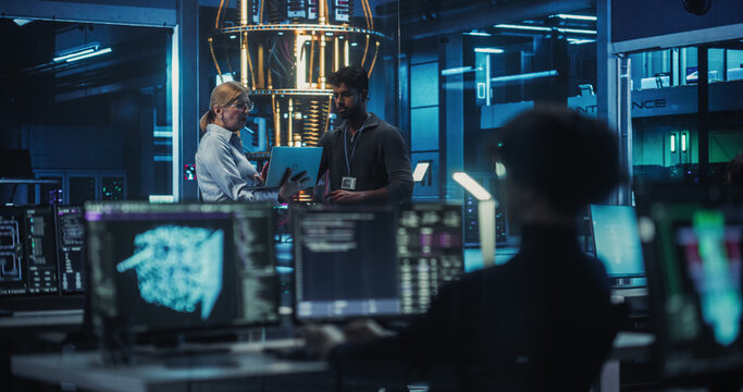 Portrait of a Middle Aged Caucasian Female Team Leader and Young Indian Engineer Talking in an Office with a Futuristic Quantum Computer Device. Specialists Discussing Research Developments - Powered by Adobe