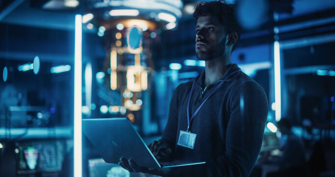 Indian Software Developer Standing in a Dark Modern Office, Working on a Laptop Computer. Young Diverse Specialist in Artificial Intelligence is Developing a Quantum Machine Learning Algorithm - Powered by Adobe