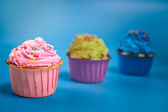 Three colorful cupcakes with pink, yellow, and blue frosting topped with sprinkles are arranged in a row on a blue background, with the pink cupcake in sharp focus up front