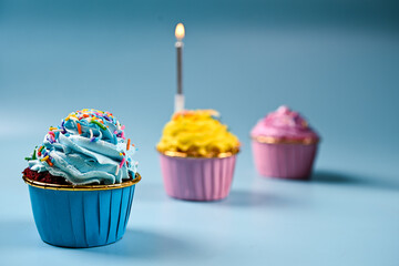 Colorful cupcakes with swirled frosting, sprinkles, and one lit candle are arranged in a soft blue background, creating a festive and joyful birthday celebration setup
