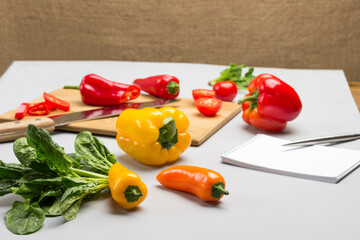 Pepper and knife on cutting board. Peppers and spinach leaves on table.  Pen and notebook