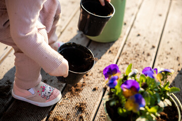 A child in light pink clothing holds a black pot on a wooden deck with soil scattered around. Purple and yellow flowers in a pot are visible in the foreground.