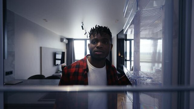 Hungry Black man opening fridge and finding empty shelves, food shortage problem