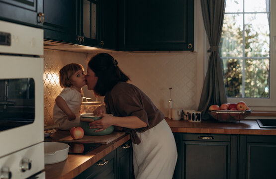 A mother kisses her young daughter in a warm kitchen filled with natural light. They are interacting near a bowl while apples are visible on the countertop, creating a homey atmosphere - Powered by Adobe