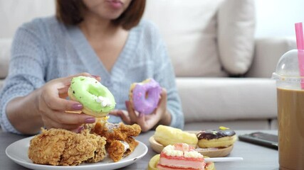 close-up of a woman’s hand holding and eating a donut, with more junk food like fried chicken, cake, sweet drinks, and desserts on the table, showing unhealthy eating habits, overeating,
