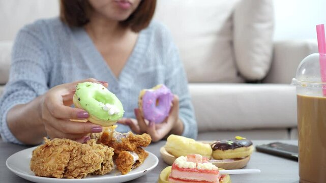 close-up of a woman&rsquo;s hand holding and eating a donut, with more junk food like fried chicken, cake, sweet drinks, and desserts on the table, showing unhealthy eating habits, overeating,