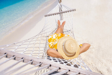 A young woman in a straw hat and a summer dress is lying in a hammock near the ocean.
