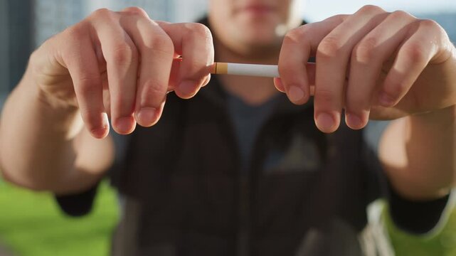 man standing with sunlit hands brought forward gripping and snapping cigarette mid fail gesture in front of blurred building and lush greenery, attempt to quit smoking