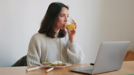 A young woman enjoys a healthy breakfast while working remotely. She eats avocado toast and drinks orange juice while checking her laptop and phone. - Powered by Adobe