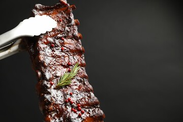 Holding delicious roasted pork ribs with tongs on black background, closeup. Space for text