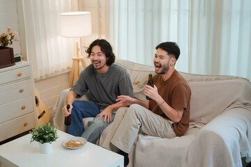 Two Asian men sit on a couch in a cozy living room, sharing laughter and drinking beer while...