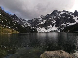 Snowy Tatra Mountains loom over the dark rippling waters of Morskie Oko lake under dramatic clouds, with rocky shoreline and forest near Zakopane, Poland