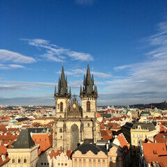 Obraz premium The Gothic spires of the Church of Our Lady before Týn rise above Prague’s historic Old Town, surrounded by colorful rooftops under a vibrant blue sky.