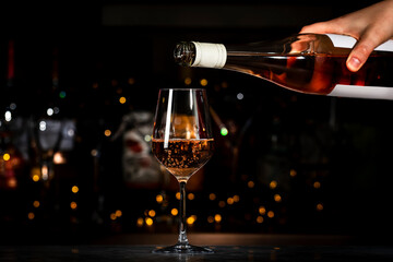 Fototapeta premium Rose pink wine glass and bottle in bartender hand on the black bar background with bokeh lights