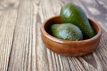 Close-up of two ripe avocados resting in a round wooden bowl placed on a rustic wood grain surface with natural light and textured background