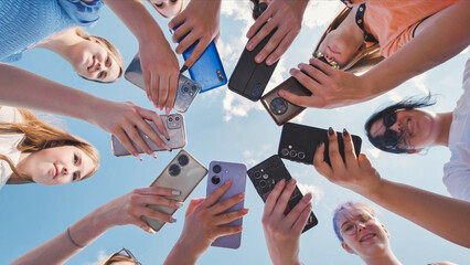School friends holding smartphones in a circle with sky background, captured from a low angle