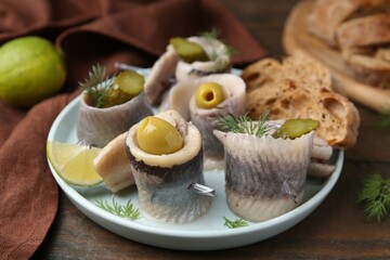 Delicious herring rolls with olives, pickles, dill and bread pieces on wooden table, closeup