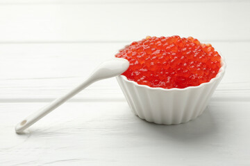 Tasty red caviar in bowl and spoon on white wooden table, closeup