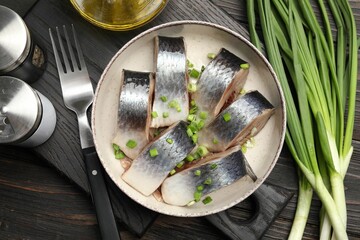 Pieces of delicious herring with green onions served on black wooden table, flat lay