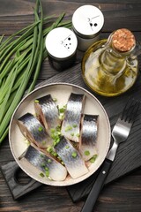 Pieces of delicious herring with green onions served on black wooden table, flat lay