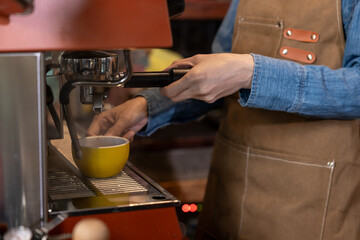 Closeup of hand preparing espresso coffee in yellow cup using machine. Casual brown apron and denim shirt reveal modern cafe setting with attention to detail in small barista business.