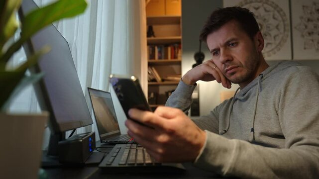 Tired freelancer procrastinating at home office, scrolling phone instead of working. Unproductive man using smartphone at home desk. Concept of procrastination and distraction in remote work