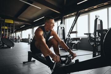 Cheerful attractive man smiling while using rowing machine in contemporary gym. Fit young man...