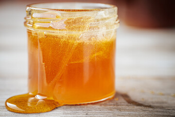 A close-up of a glass jar filled with golden honey spilling over the sides onto a rustic wooden surface. The honey shimmers in rich amber hues.