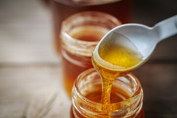 A close-up of golden honey slowly dripping from a plastic spoon into a glass jar, with more jars in the background softly blurred on a rustic wooden surface.