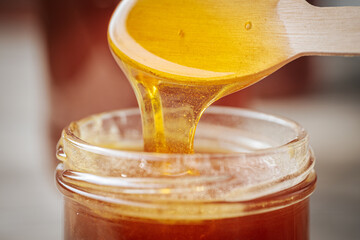 A close-up of golden honey dripping from a wooden spoon into a glass jar, showcasing its rich,...