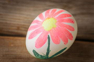 A white egg decorated with a hand-painted pink flower and green stem rests on a wooden surface. Bright and cheerful, with a spring feel.